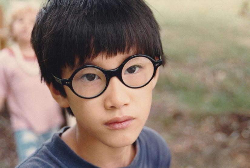 Moses Farrow as a boy, in round black-rimmed glasses, looking straight at the camera outdoors.