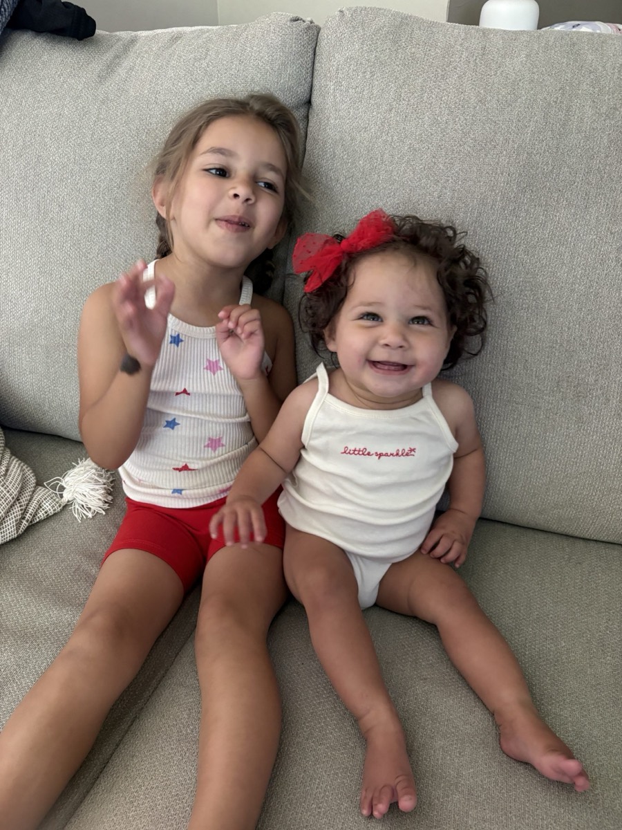 Clara Stanford and her older sister sit together on a sofa, both smiling at the camera.