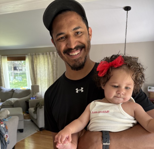 Louis Stanford, wearing a black cap and T-shirt, smiles as he holds his baby daughter Clara, who wears a red hair bow, in the family's living room.