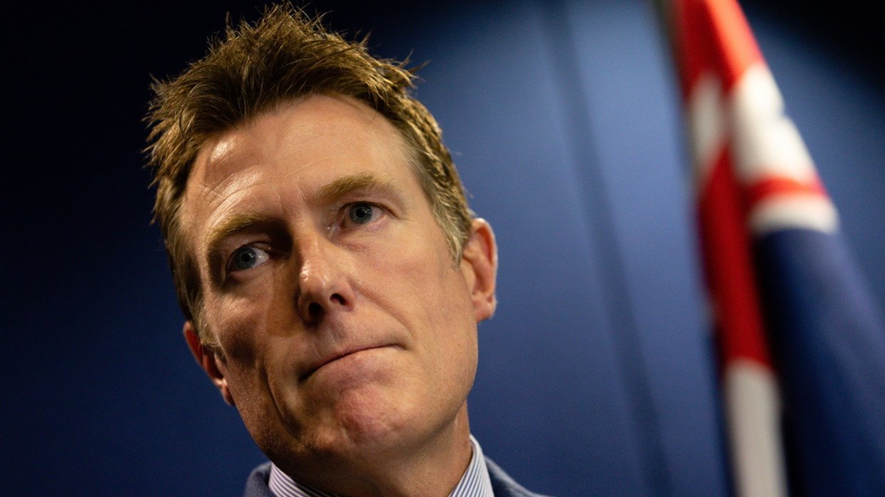 Close-up of Christian Porter at a press conference, tight-lipped, with an Australian flag blurred behind him.