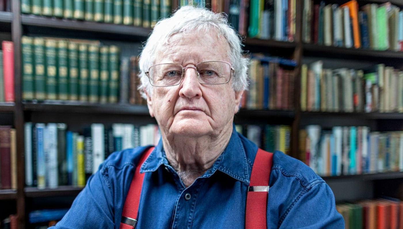Campbell Malone, white-haired and wearing rimless glasses, a denim shirt and red braces, sits in front of packed bookshelves.