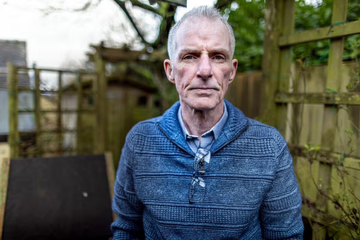 Eddie Gilfoyle, clean-shaven and grey-haired in a blue knitted jumper with reading glasses hooked at the collar, stands in a garden beside a wooden fence.