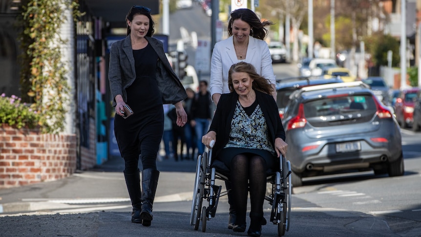 Sue Neill-Fraser in a wheelchair on a footpath, accompanied by her two daughters.