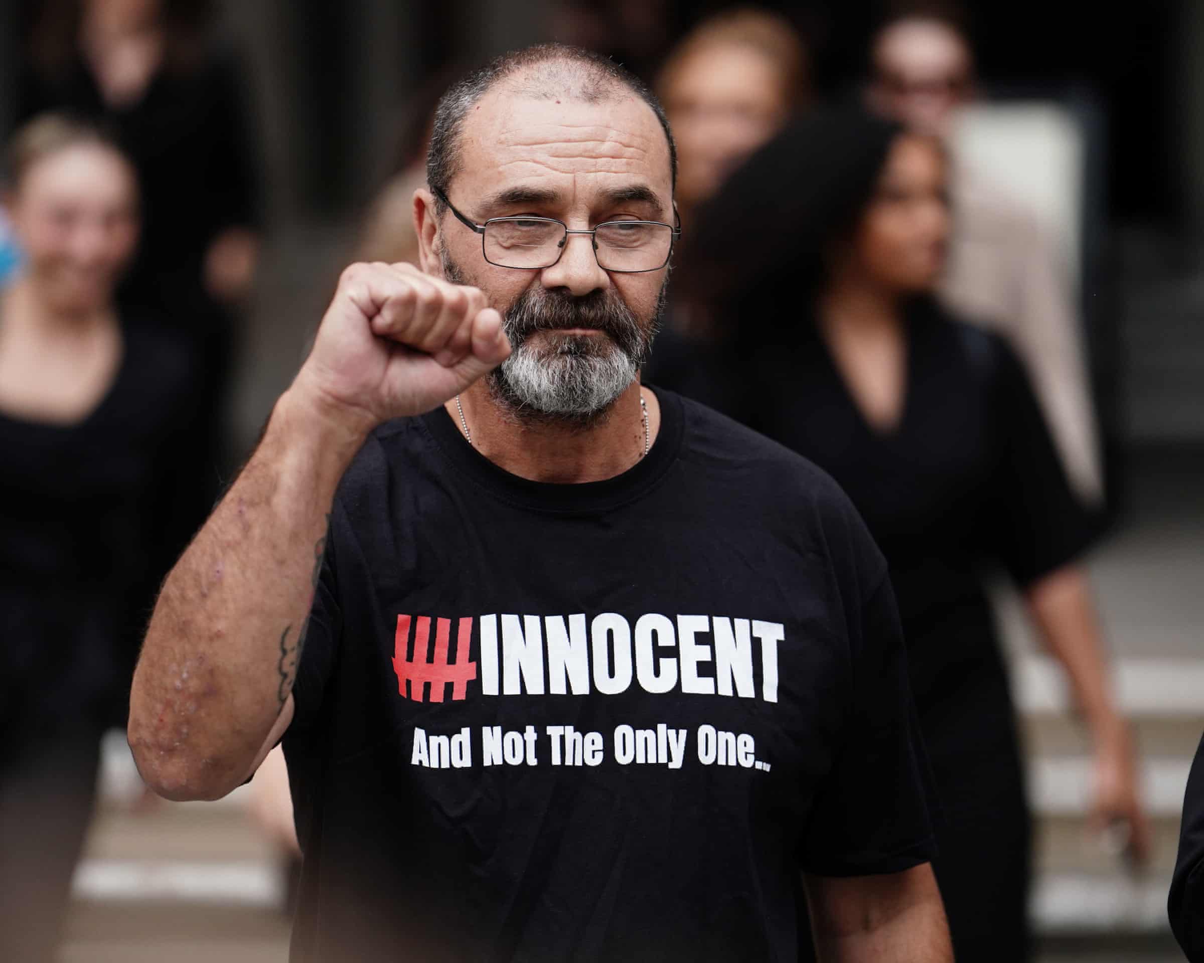 Andrew Malkinson, bearded and wearing glasses and a black T-shirt reading “Innocent — And Not The Only One”, raises a clenched fist as he leaves court.