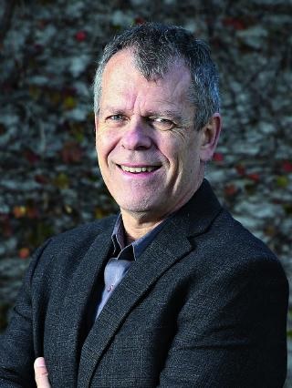 Portrait of Professor Richard Bryant, arms folded, in a dark jacket and open-necked shirt in front of a stone wall.